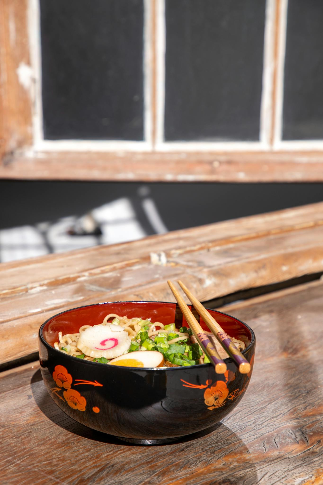 Black ceramic bowl of ramen with chopsticks set on wooden table, natural light streaming through a window.